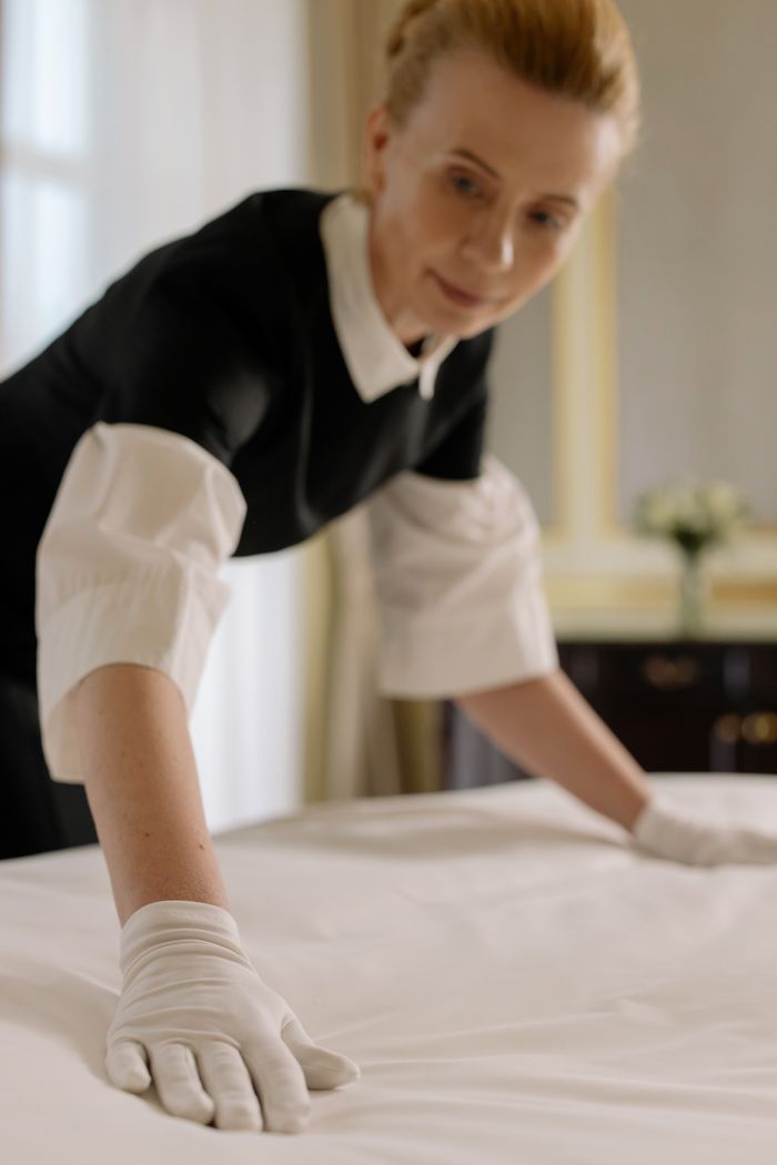 services-04 A housekeeper elegantly arranges bed sheets in a luxurious hotel room, showcasing hospitality expertise.